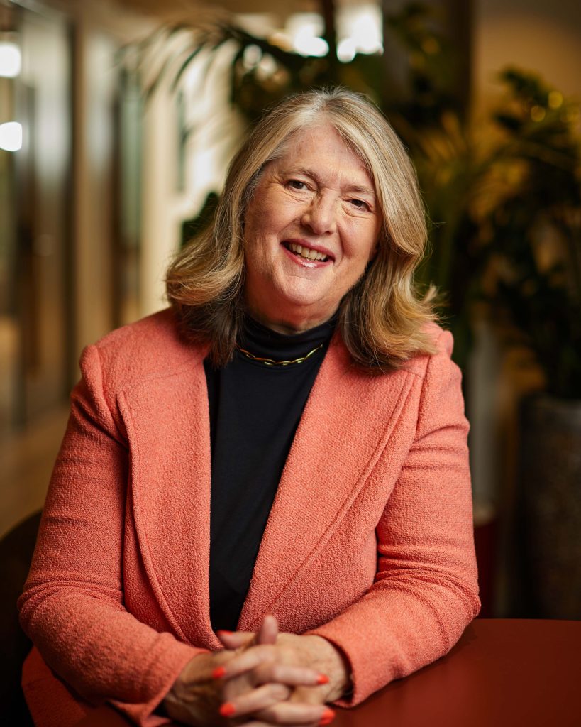 A portrait photo of Anne Glover in an open plan office setting, wearing a bright blazer and dark top. She is smiling at the camera while sat at a table with her hands together, resting on the tabletop.