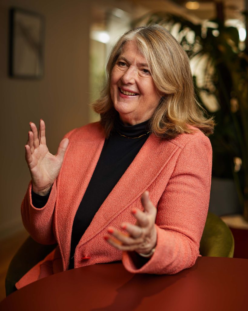 A portrait photo of Anne Glover in an open plan office setting, wearing a bright blazer and dark top. She is smiling and looking away from the camera with her hands in the air gesturing in front of her.