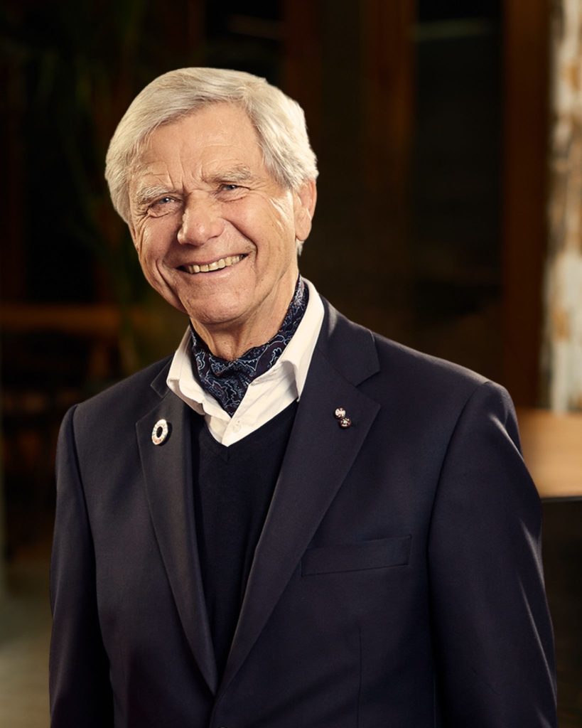 A portrait photo of Hermann Hauser in a relaxed office setting. Hermann is smiling and is wearing a blazer, shirt and a cravat around his neck.