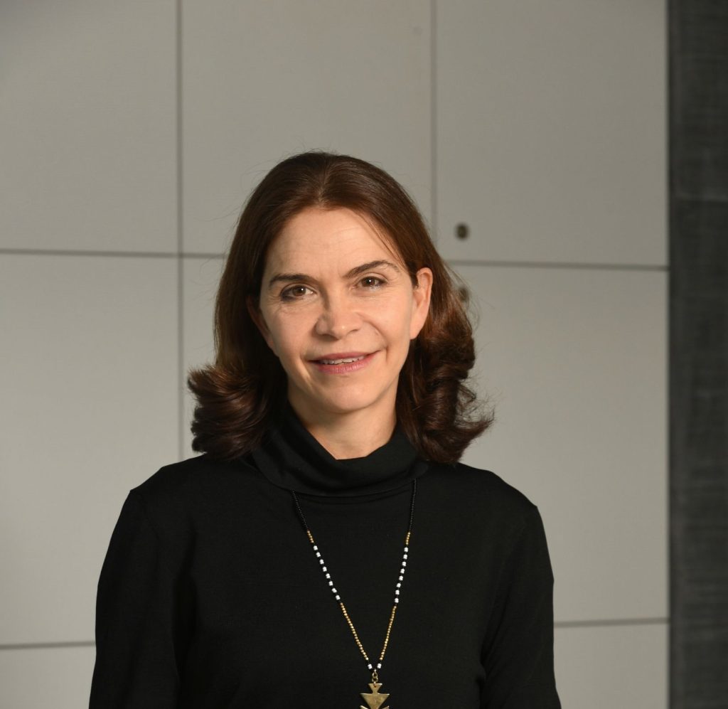 A portrait photo of Kelly Richdale at the exterior of a building with square plain cladding to be seen in the background. Kelly is looking at the camera directly and smiling. She is wearing a dark turtle shawl neck top with a long necklace.