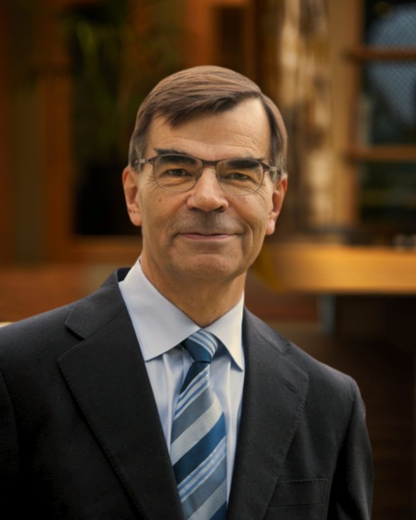 A portrait of Professor Steve Young in an open plan office setting for Amadeus Capital Partners. Steve is wearing a dark blue suit and light blue shirt with striped tie. He is smiling at the camera.