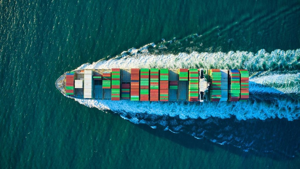 An aerial view of a container ship sailing through the ocean with an array of containers in different colours on its deck. The image represents the global supply chain network which Altana AI has mapped.