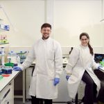 Two male and female Constructive Bio staff members wearing white lab coats and gloves smiling for a photo together in the laboratory,