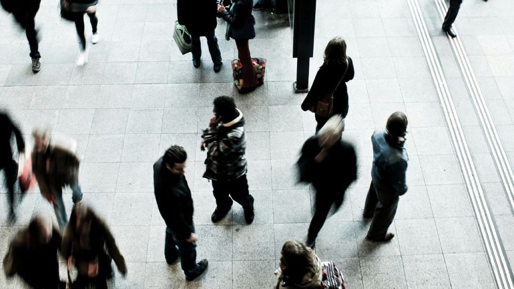 A group of people walking around a train station, most are blurred and unclear, with one individual in focus in the centre of the photo who is using their phone.