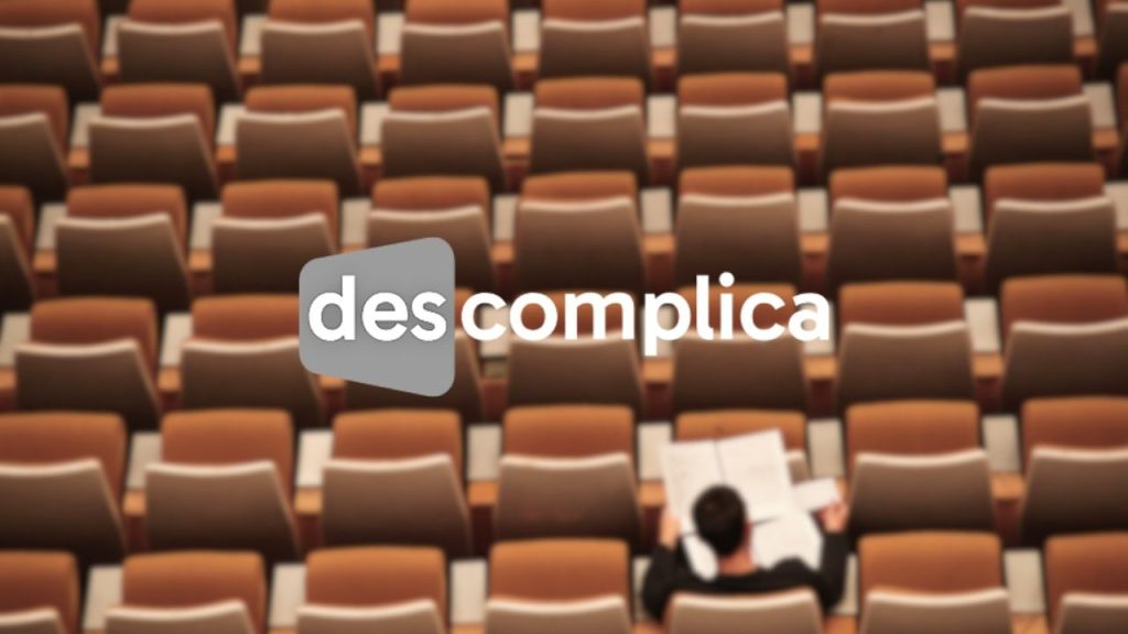 Descomplica company logo overlaid on a photo from above a a lecture theatre seating area with a single student sat in one of the seats, studying some papers.