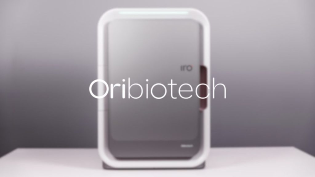 Oribiotech company logo overlaid on a photo of the IRO manufacturing platform which enables advanced cell and gene therapy for applications like cancer treatment. The device is rectangular, like a small desktop fridge, with the letters "IRO" printed on the door.