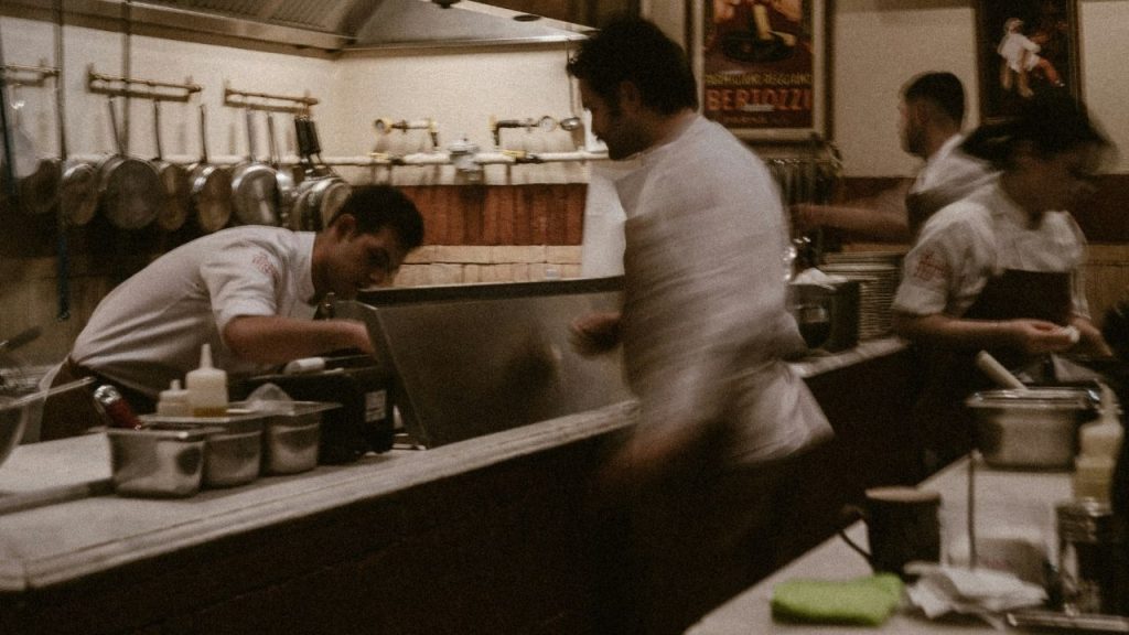 a restaurant scene showing a chef and cooking line alongside the front of house waiting staff to represent Tenzo's restaurant operations management platform.