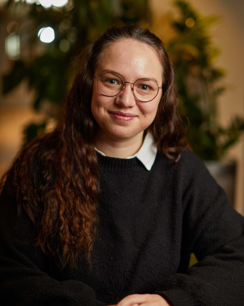 A portrait photo of Elsa Roudot sitting in an open plan office setting. Elsa is smiling at the camera and wearing a dark jumper over an oxford shirt with hair falling to one side in front of her shoulder. Behind Elsa is a plant with leaves.