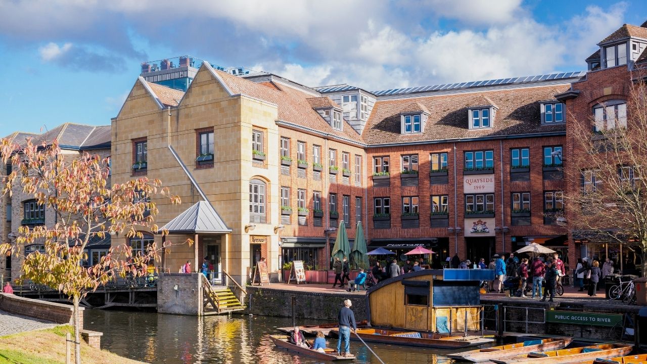 A photo of the Amadeus Capital Partners Cambridge Quayside office which also includes the river with a punting boat and passengers aboard, and a group of people congregating outside restaurants in the Quayside courtyard.