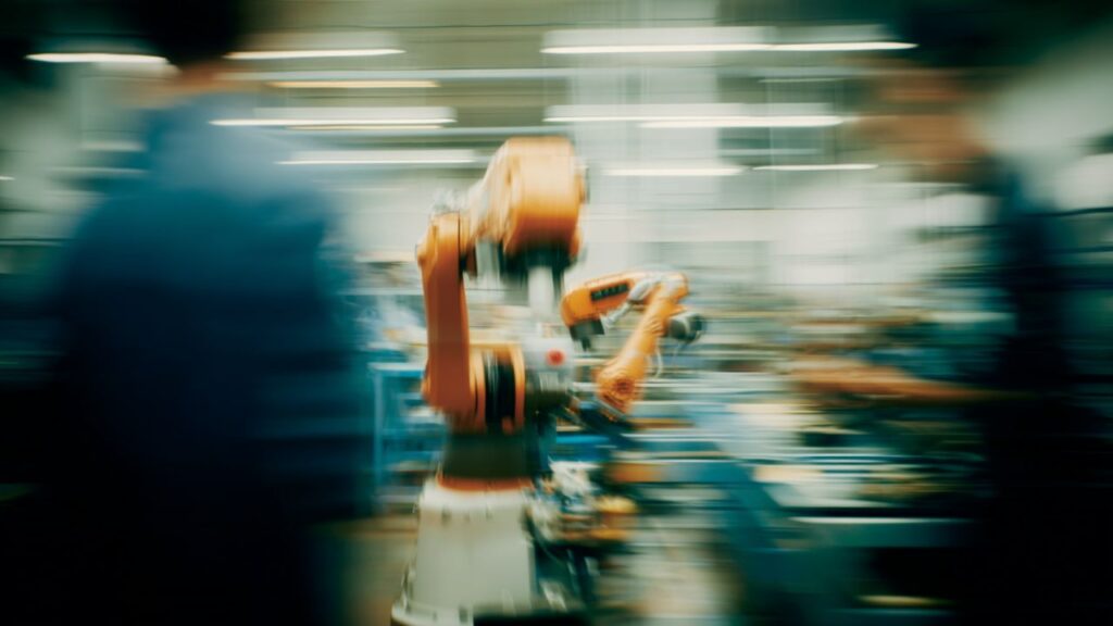 A set of robotic arms in a factory setting with humans present.